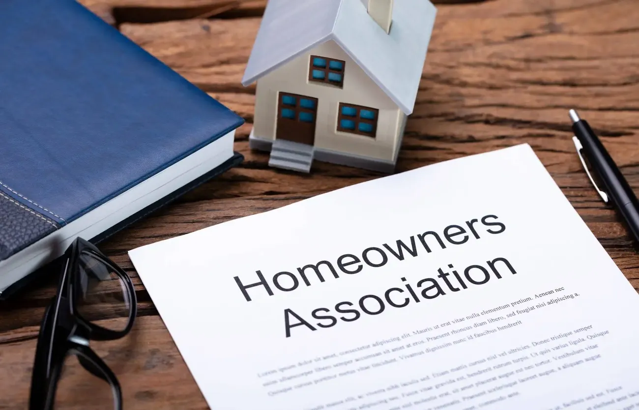 Paper labeled 'Homeowners Association' next to a model house, glasses, book, and pen on a wooden table.